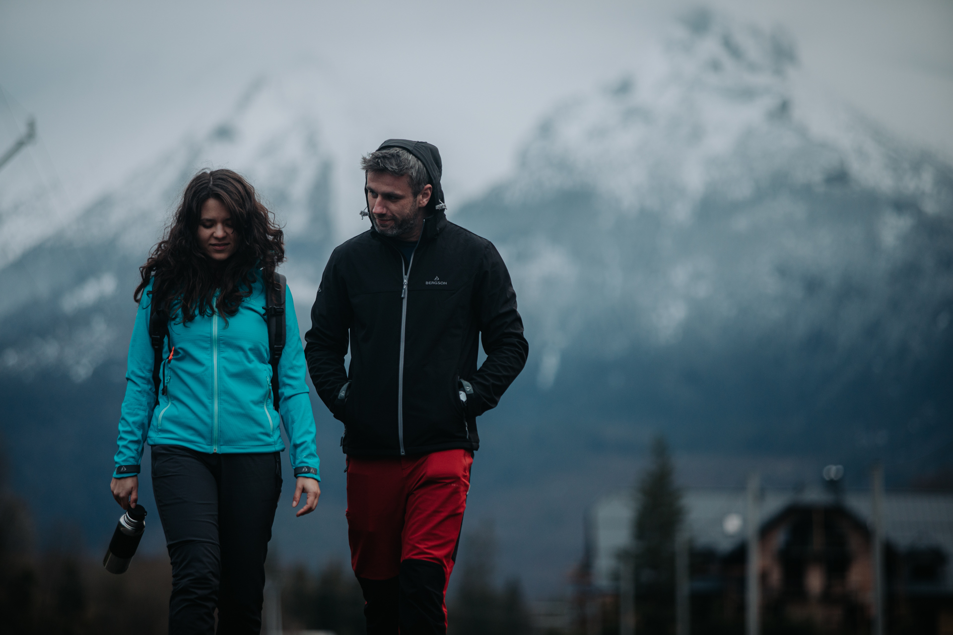 Kobieta w turkusowej kurtce i czarnych spodniach z plecakiem, mężczyzna w czarnej kurtce i czerwonych spodniach; w tle górski krajobraz.