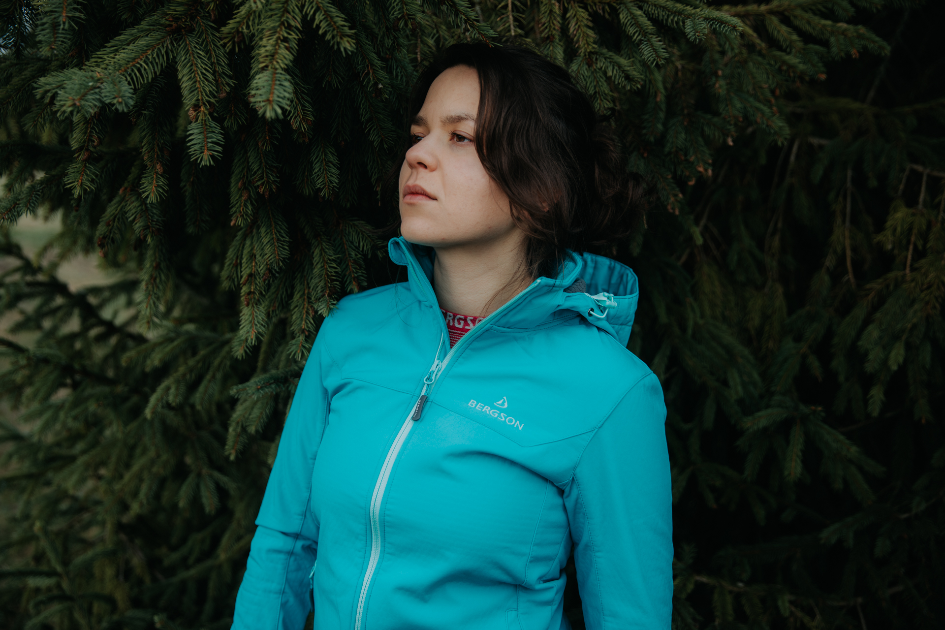 Szatynka z brązowymi oczami patrząca w lewo, w turkusowej kurtce Bergson, stojąca na tle drzewa iglastego.